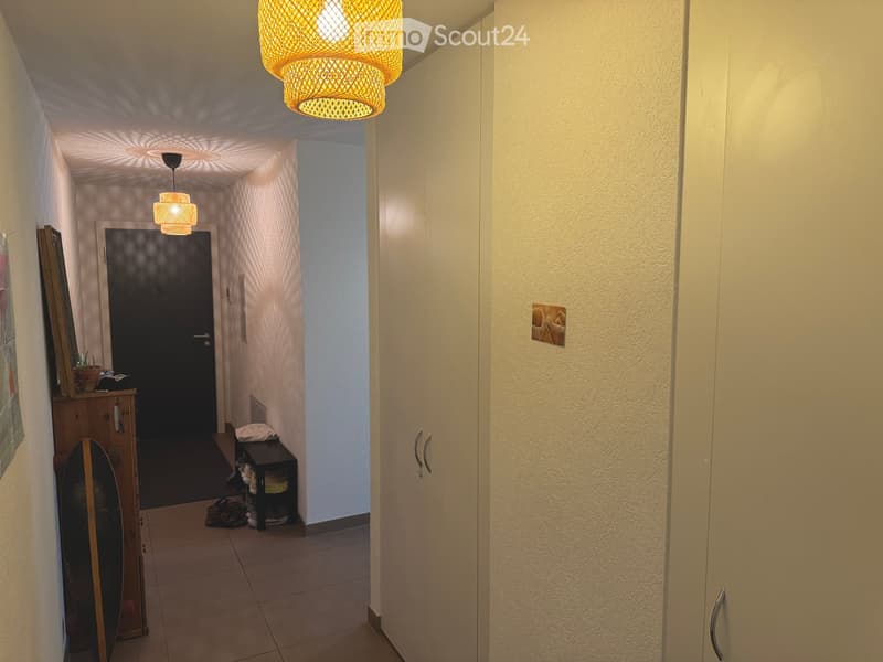 A narrow hallway with beige walls, tiled floor, and two hanging pendant lights. There is a small wooden cabinet with a plant and a shelf with shoes and a folded towel. A closed door is visible at the end of the hallway.