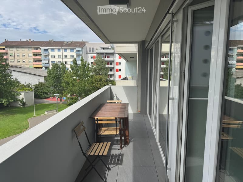 Balcony with a wooden table and a folding chair, overlooking a green area with trees and buildings.
