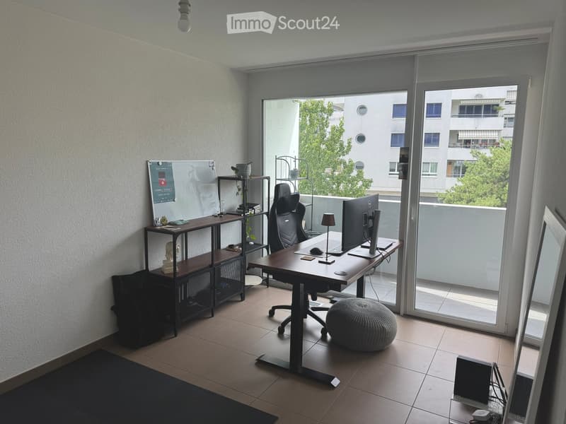 Office space with a desk, chair, laptop, screen, stool, shelf, and lamp. Large window with a view of the building outside.