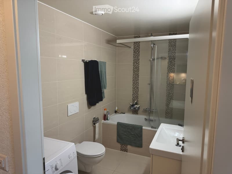 Modern bathroom with beige tiles, a bathtub with a glass shower enclosure, a toilet, a sink, a washing machine, and towels.