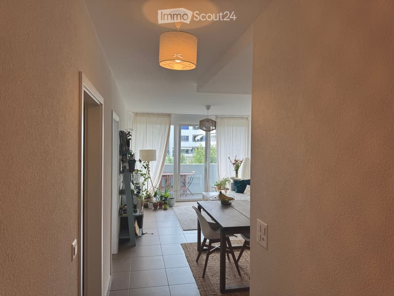 A modern, open-plan living and dining area with a large window and sliding glass door leading to a balcony. The space features a dining table with chairs, a sofa, potted plants, and a floor lamp.