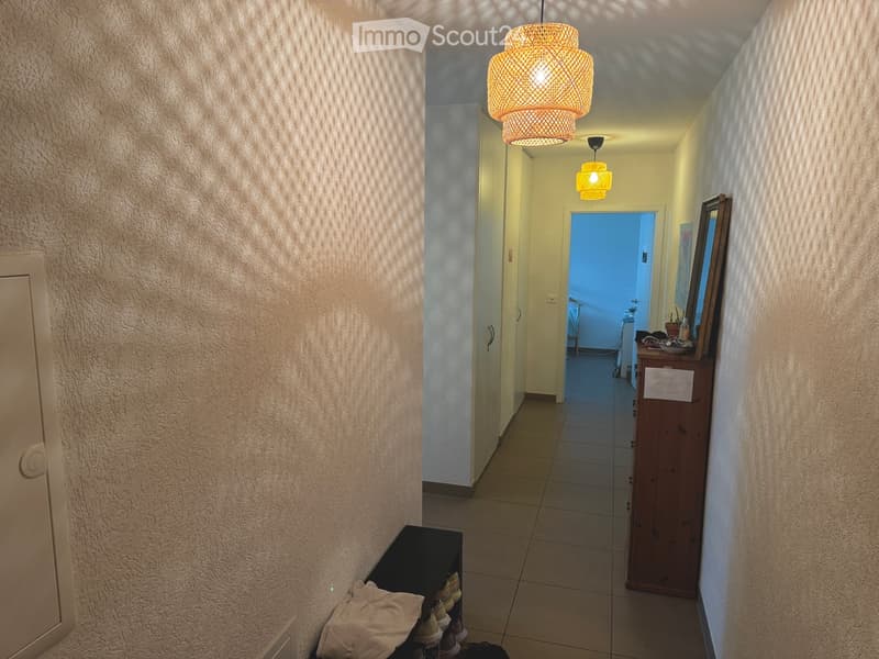 A well-lit hallway with beige walls, tiled floor, and a hanging woven light fixture. A small wooden cabinet and a mirror are visible on the right side, with a partially open door leading to another room.