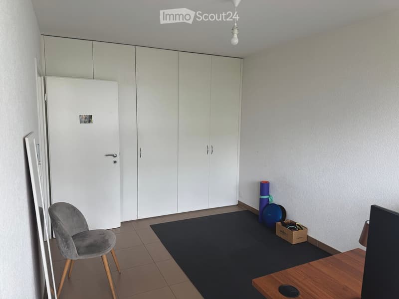 A modern, minimalistic room with a grey upholstered chair, a wooden table with a screen, a large mirror, and white storage cabinets.