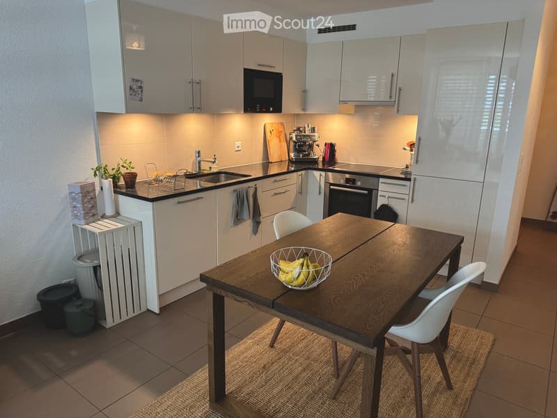 Modern kitchen with white cabinets, stainless steel appliances, a wooden dining table with chairs, and a bowl of bananas on the table.
