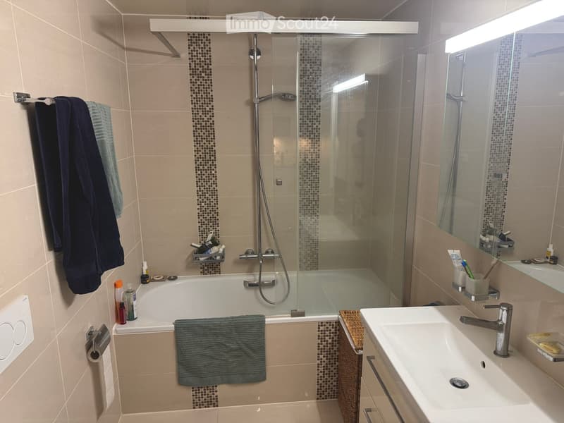 Modern bathroom with a bathtub, glass-enclosed shower, sink, mirror, and various toiletries. Beige tiles and a wicker basket are visible.