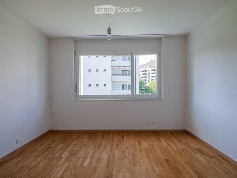 Empty room with wooden flooring, white walls, and a large window with a view of neighboring buildings.