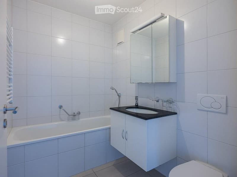 Modern bathroom with white tiled walls and floor, featuring a bathtub, a sink with a modern faucet, a mirrored cabinet, and a toilet.