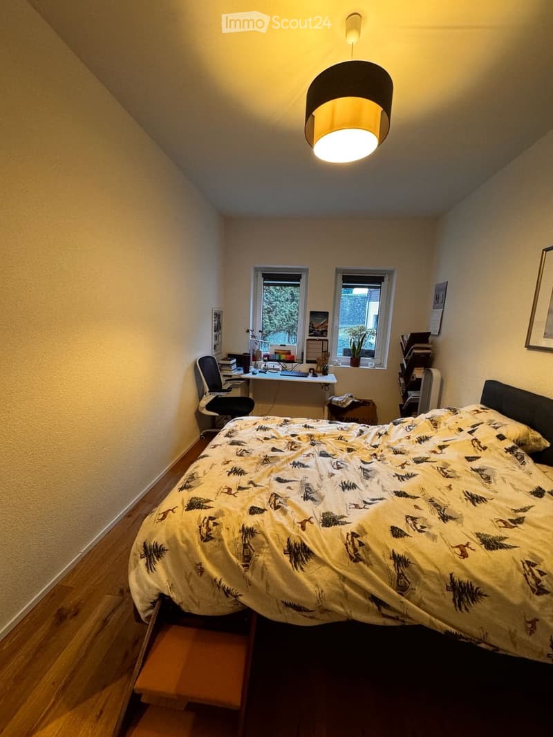 Cozy bedroom with a bed, desk, chair, and a hanging lamp. The room features wooden flooring and two windows with a view of greenery.