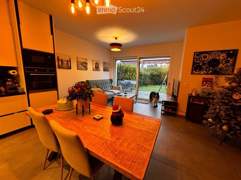 A cozy dining and living area with a wooden dining table, chairs, a sofa, a Christmas tree, and a view of the garden through large windows.