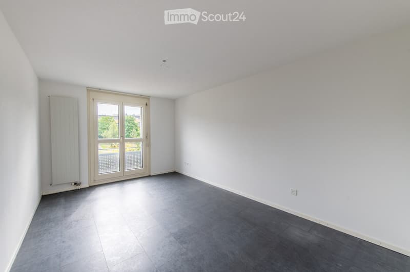Empty room with grey tiled floor, white walls, and a large window with a view of greenery outside.