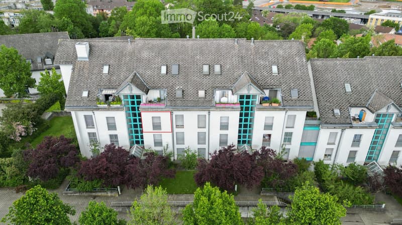A multi-story residential building with a grey tiled roof, white walls, and multiple windows. The building features balconies with green railings and is surrounded by greenery and trees.