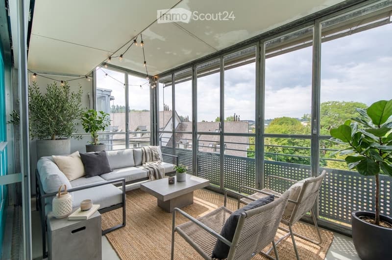 A modern, well-lit balcony with large glass windows offering a scenic view. Features a white sofa, coffee table, wicker chairs, and several potted plants.