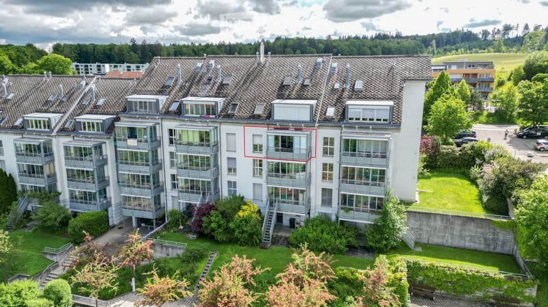 A multi-story residential building with a tiled roof, large windows, and balconies. The building is surrounded by greenery and has a well-maintained lawn and pathways.