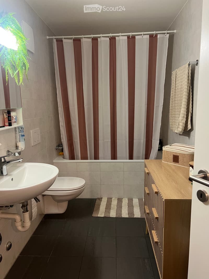 Modern bathroom with a sink, toilet, bathtub, shower curtain, towel, wooden cabinet, and mirror. The space is well-lit with natural light from a window on the left side.