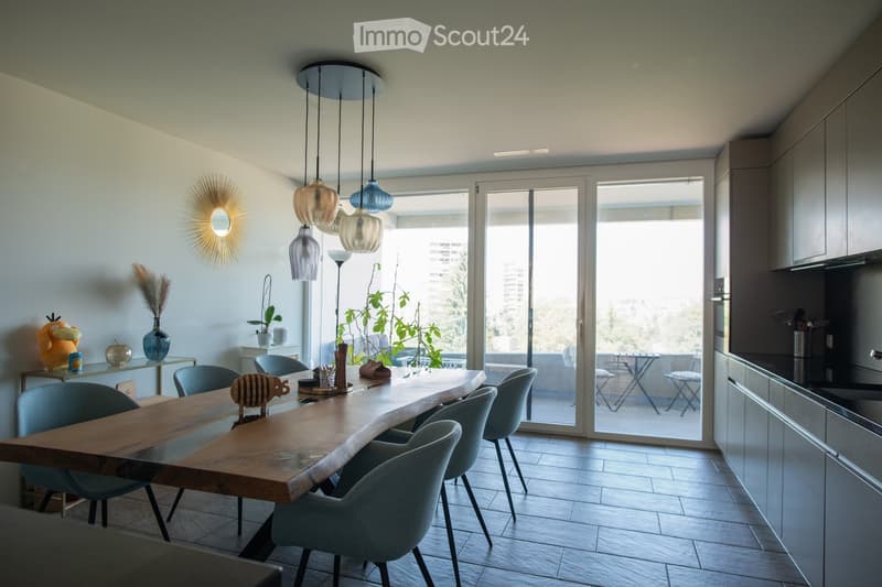 Modern kitchen and dining area with a long wooden table, grey chairs, pendant lights, and a view of a balcony with outdoor furniture.