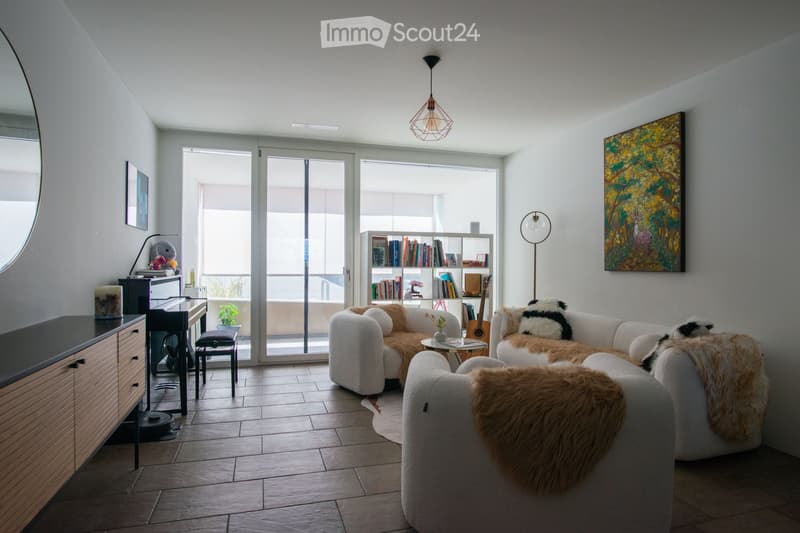 Modern living room with large sliding glass doors leading to a balcony, white sectional sofa with fur throws, coffee table, bookshelf, guitar, and a painting on the wall.