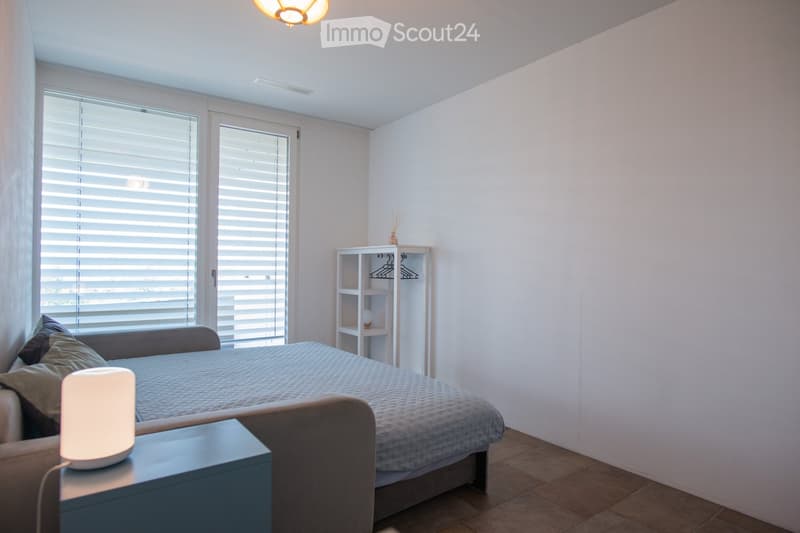 Modern bedroom with a large bed, nightstand with lamp, small shelf, and large windows with blinds.