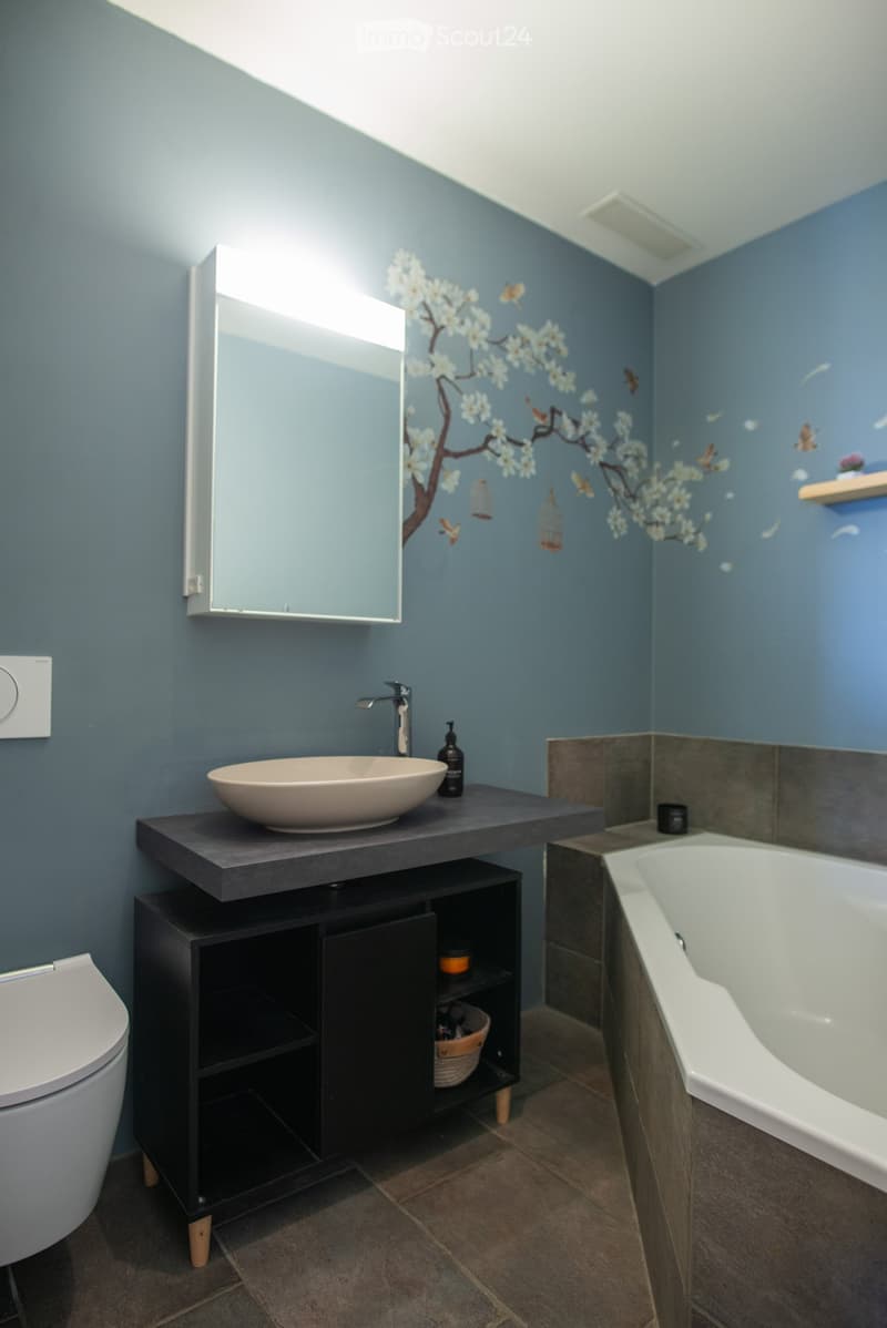 Modern bathroom with dark blue walls, a large mirror, a sleek black countertop sink, a bathtub, and a toilet. The decor includes a floral mural and wooden shelves.
