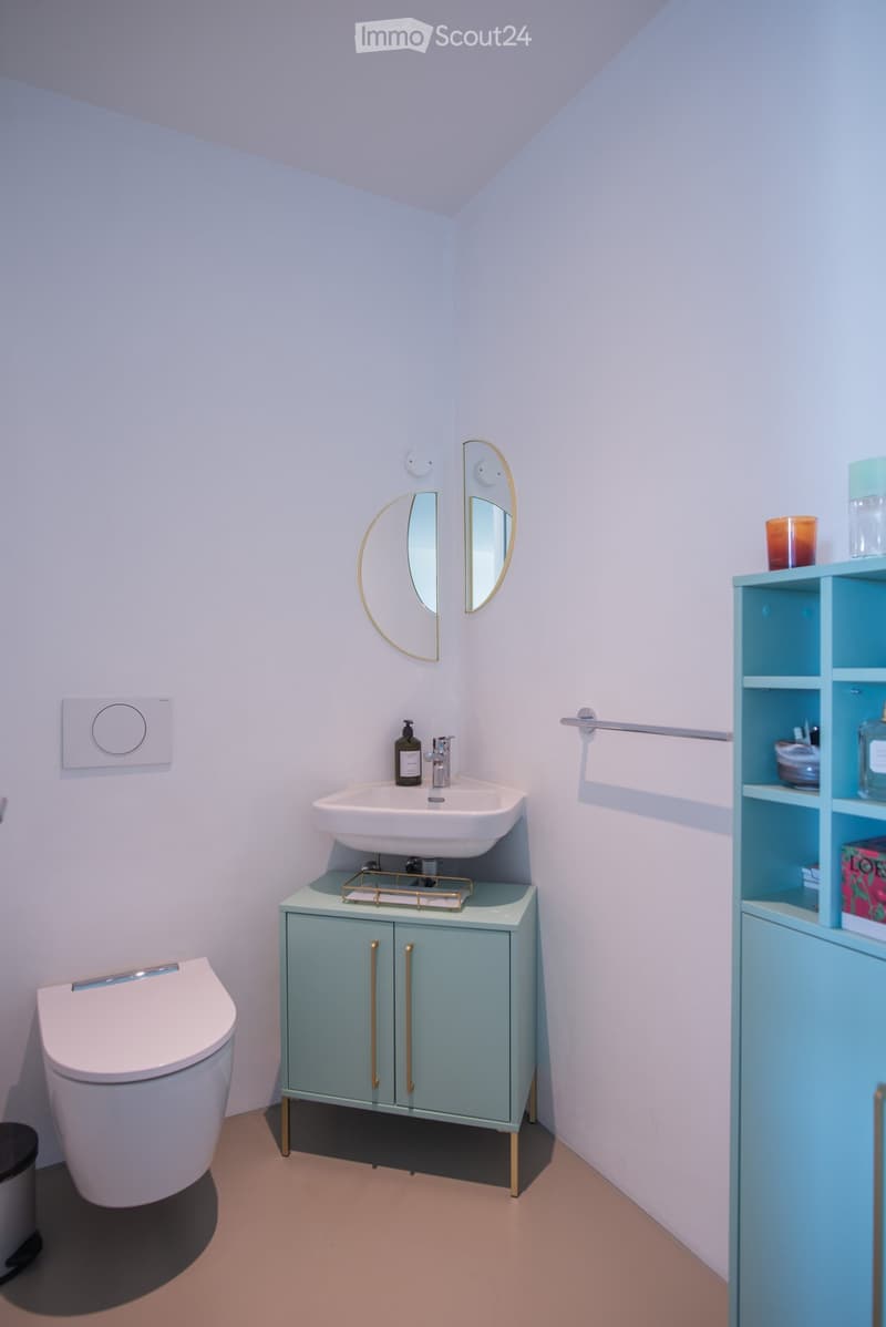 Modern bathroom with a white toilet, a light blue cabinet with gold handles, a sink with a mirror, and a towel rack. The walls are light blue, and the floor is light beige.