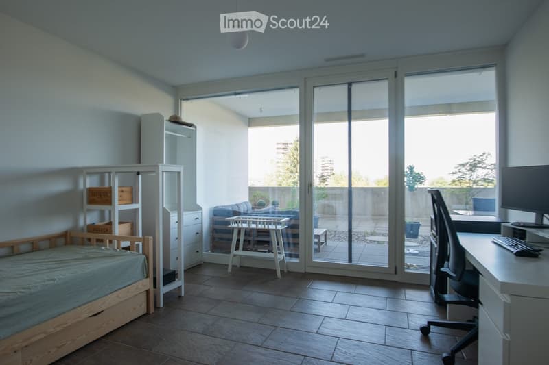 A modern, well-lit room with a bed, desk, chair, and a sliding glass door leading to a small outdoor area. The room features a tiled floor, white walls, and a mix of wooden and white furniture.