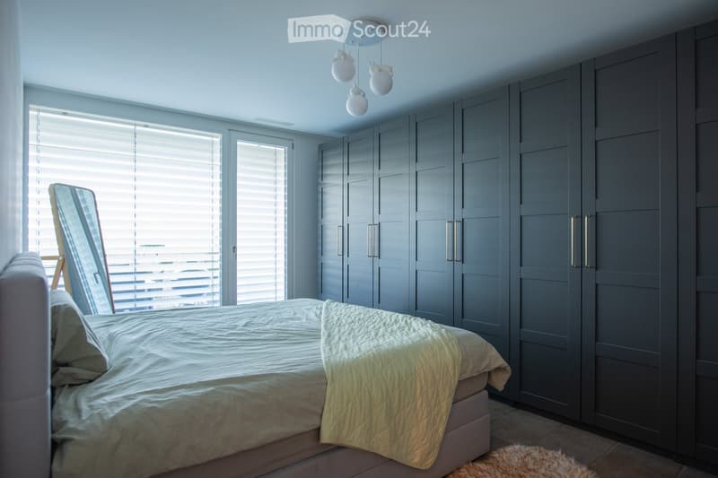 Modern bedroom with a large bed, grey wardrobe, large windows with blinds, and a mirror leaning against the wall.
