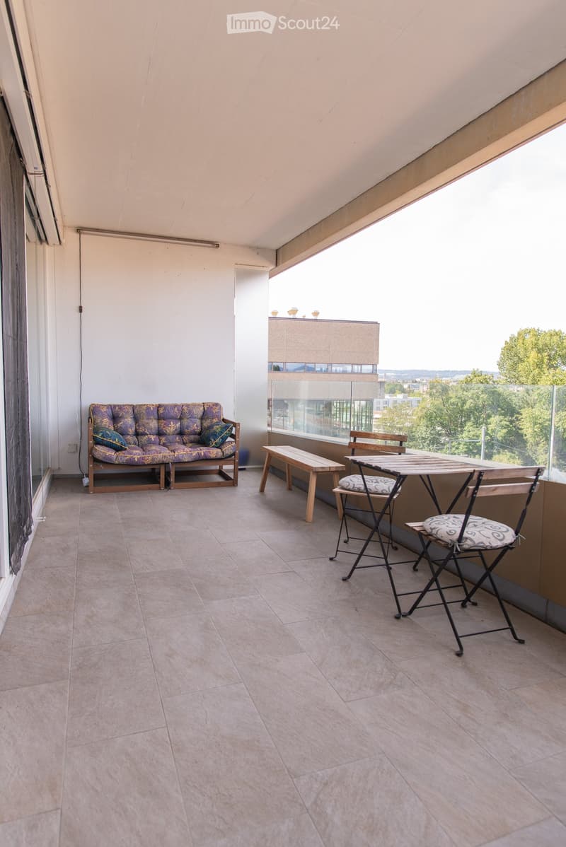 Balcony with tiled floor, sofa, table, chairs, and bench, overlooking a green landscape.