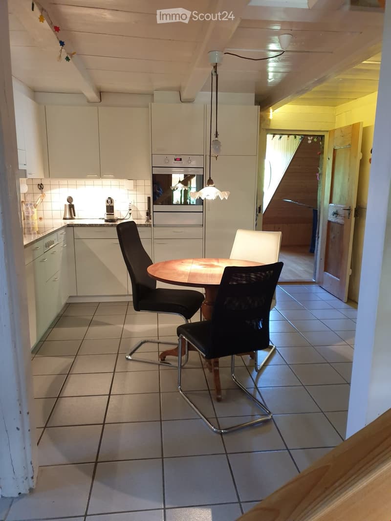 Bright kitchen with white cabinets, a round wooden dining table with four chairs, and a hanging lamp. The kitchen features tiled floors and a visible doorway leading to another room.