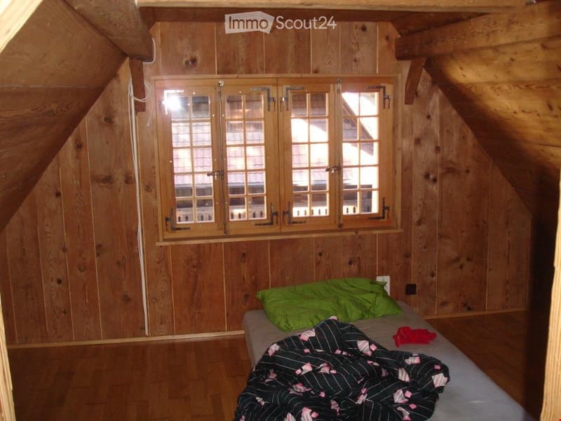 Cozy attic bedroom with wooden walls and slanted ceilings, featuring a single bed with a green pillow and a patterned blanket, and a window with wooden shutters.