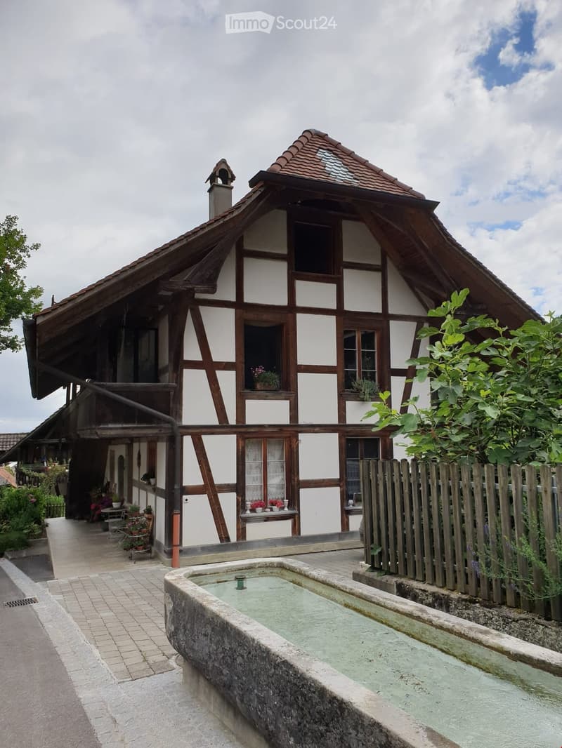 Traditional half-timbered house with white plaster walls, wooden beams, and a tiled roof. Features a small porch with a table and chairs, a wooden fence, and a stone basin in the foreground.