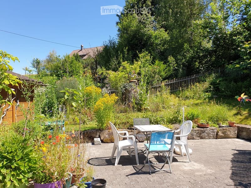 Garden patio with a table and chairs, surrounded by lush greenery and potted plants, with a stone wall and a wooden shed in the background.