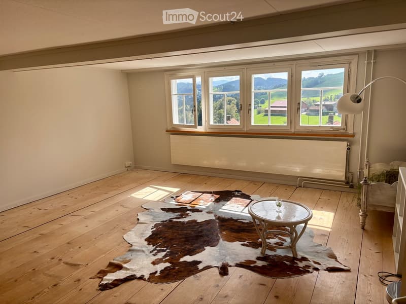 Bright, unfurnished room with wooden flooring, large windows offering scenic views, a small round table with a glass, and a cowhide rug.