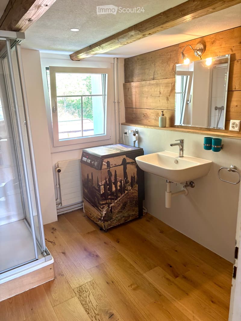 Bathroom with wooden flooring, a white sink with a modern faucet, a large mirror, a small refrigerator with a scenic design, and a window offering a view of the garden.