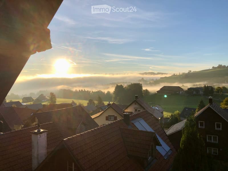 A panoramic view of a village with traditional houses and red-tiled roofs, surrounded by misty fields and hills under a clear blue sky at sunrise.