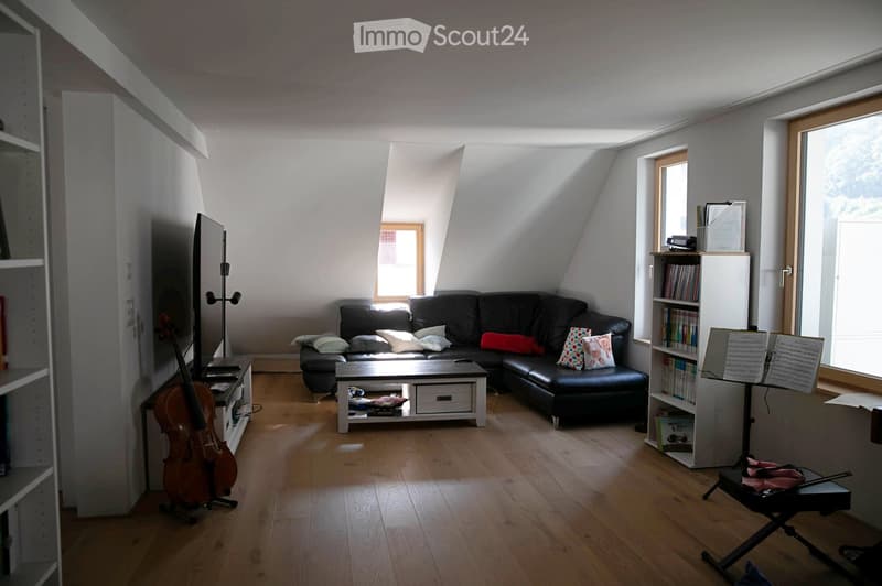 Modern living room with wooden flooring, large windows, a black sectional sofa with colorful cushions, a white coffee table, a cello, a bookcase, and a music stand.