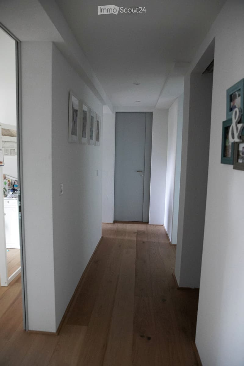 A narrow hallway with light-colored walls, wooden flooring, and a closed door at the end. There are framed pictures on the left wall and a partially visible room on the left side.