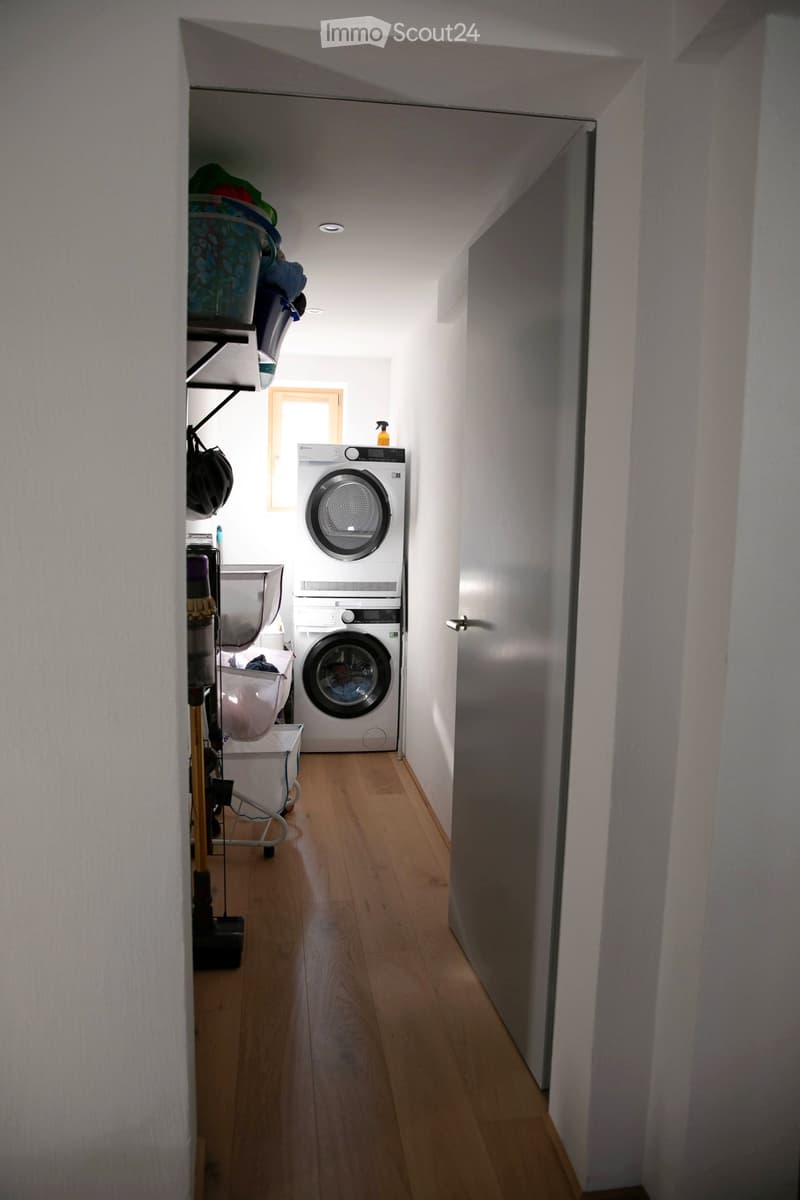 Narrow hallway with wooden flooring, white walls, and a small window. A washing machine and dryer are placed against the wall, with shelves and a stroller nearby.