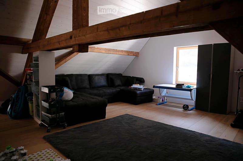 Attic living space with wooden beams, slanted ceiling, large window, black sectional sofa, coffee table, small table with items, and storage unit.