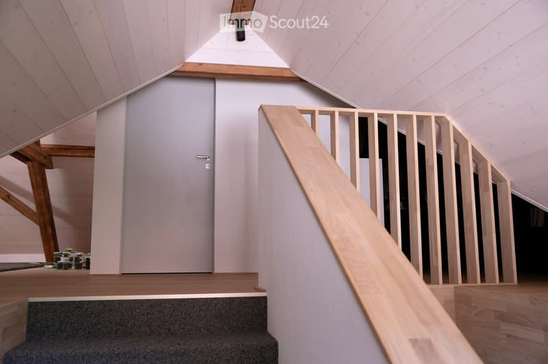 Modern hallway with wooden staircase, grey walls, and a closed door. The ceiling has exposed wooden beams and a skylight.