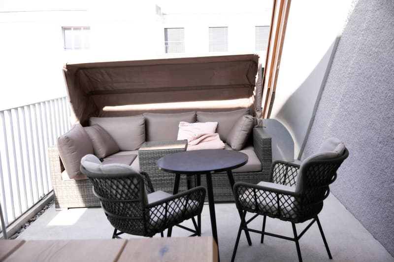 Balcony with a wicker sofa, cushions, a small round table, and two wicker chairs.