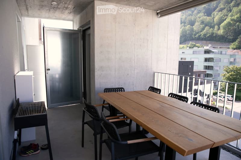 Balcony with a wooden table and black chairs, a grill, and a view of the surrounding area.