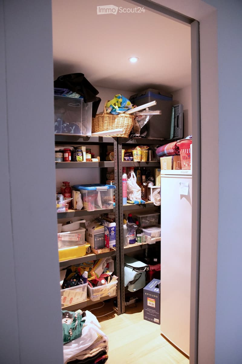 A well-organized pantry or storage room with metal shelving units filled with various household items, including food, cleaning supplies, and storage bins. A refrigerator is visible on the right side, and a basket sits on the top shelf.