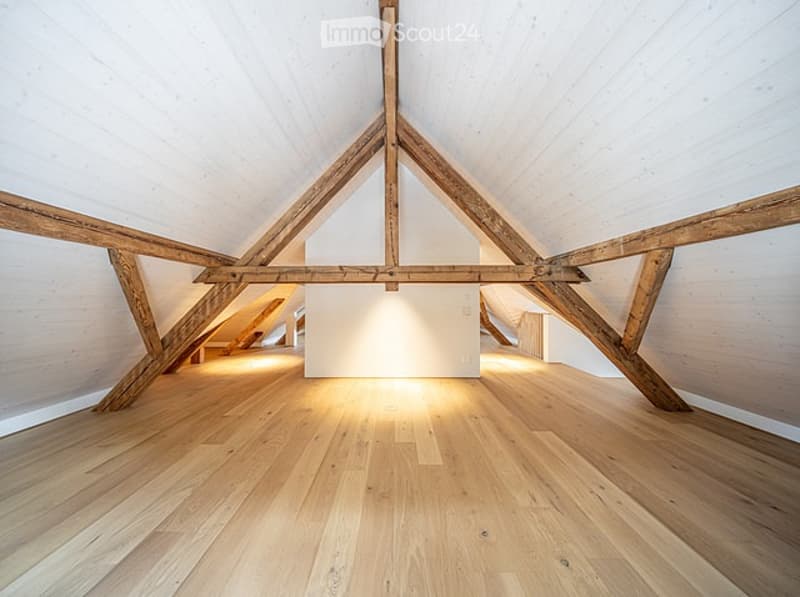 Open, airy attic space with exposed wooden beams and polished wooden flooring. The room is completely unfurnished, with white walls and ample natural light streaming in from the windows.