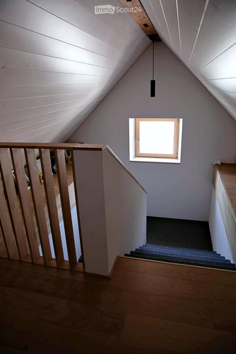 Staircase with wooden railings, grey carpeted steps, and a skylight window above, leading to an attic space with exposed wooden beams and a hanging light fixture.