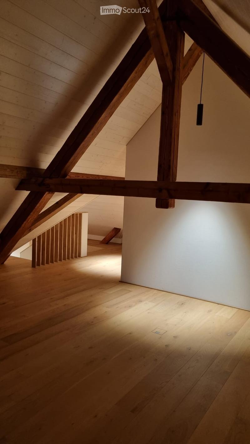 An empty attic space with wooden beams and flooring, featuring exposed wooden beams and a small staircase leading to another area.