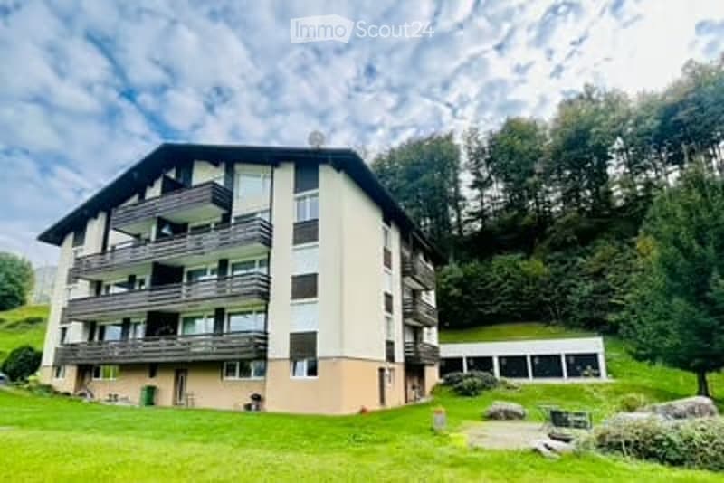 Multi-story residential building with balconies, surrounded by green lawn and trees.