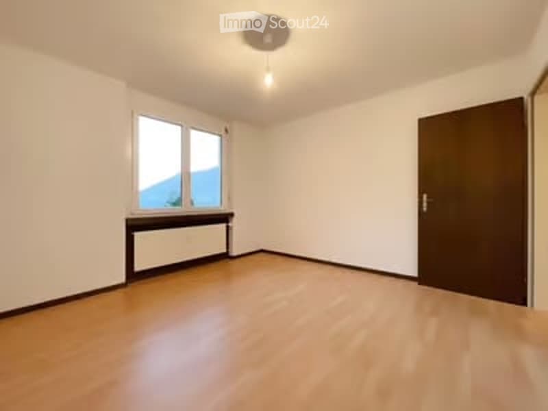 Empty room with wooden flooring, white walls, large window with radiator, and a closed door.