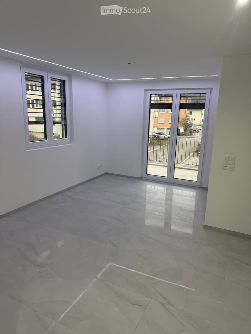 Empty room with light grey walls, polished marble floor, large window with blinds, and sliding glass door leading to a balcony.