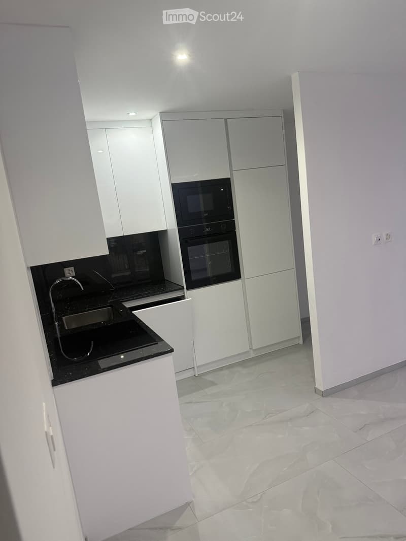 Modern kitchen with white cabinets, black countertops, a sink, faucet, oven, and microwave. The floor is tiled with a marble-like pattern.