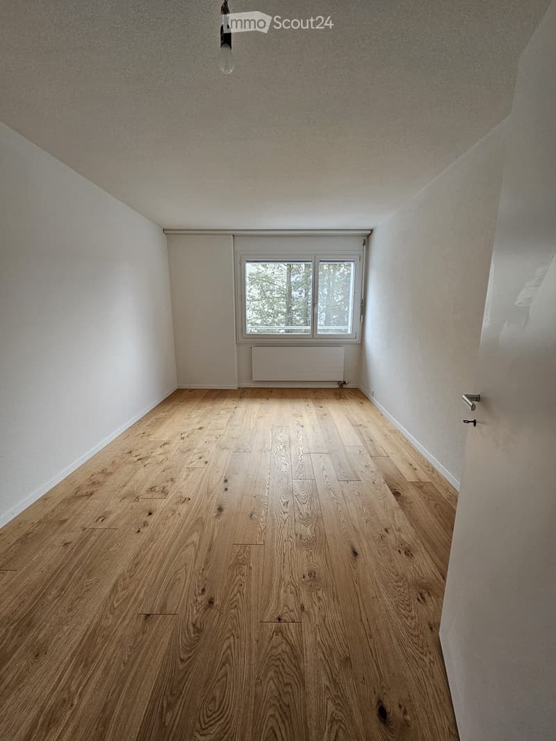 Empty room with light wooden flooring, white walls, and a window with a radiator below it. The room has a simple, clean design with no furniture or decorations.