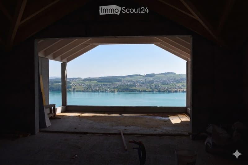 Empty room with exposed wooden beams, large window offering a scenic view of a lake and surrounding landscape.